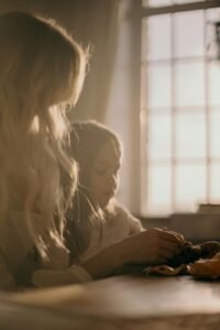 A warm, intimate moment between a mother and daughter sitting by a sunlit window indoors.